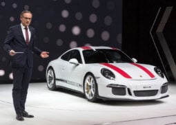Man in suit stands by Porsche in showroom.