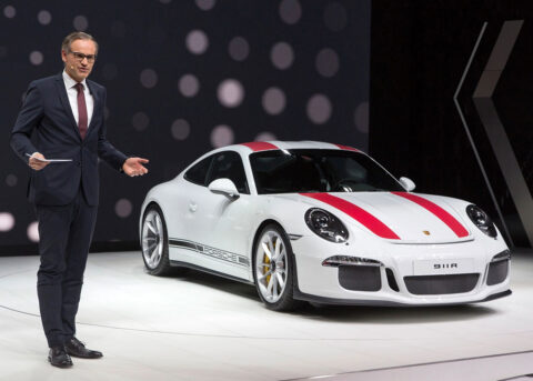 Man in suit stands by Porsche in showroom.