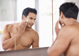 Shirtless man applies face cream in modern bathroom.