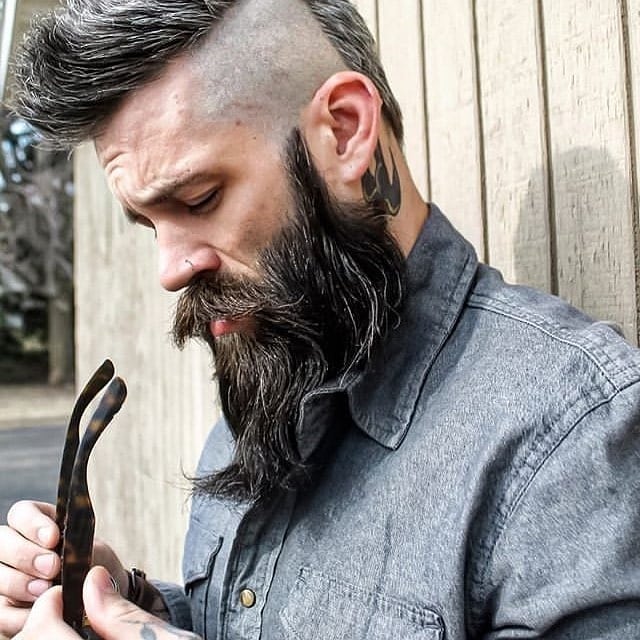 Bearded man examines wood, wears denim shirt.