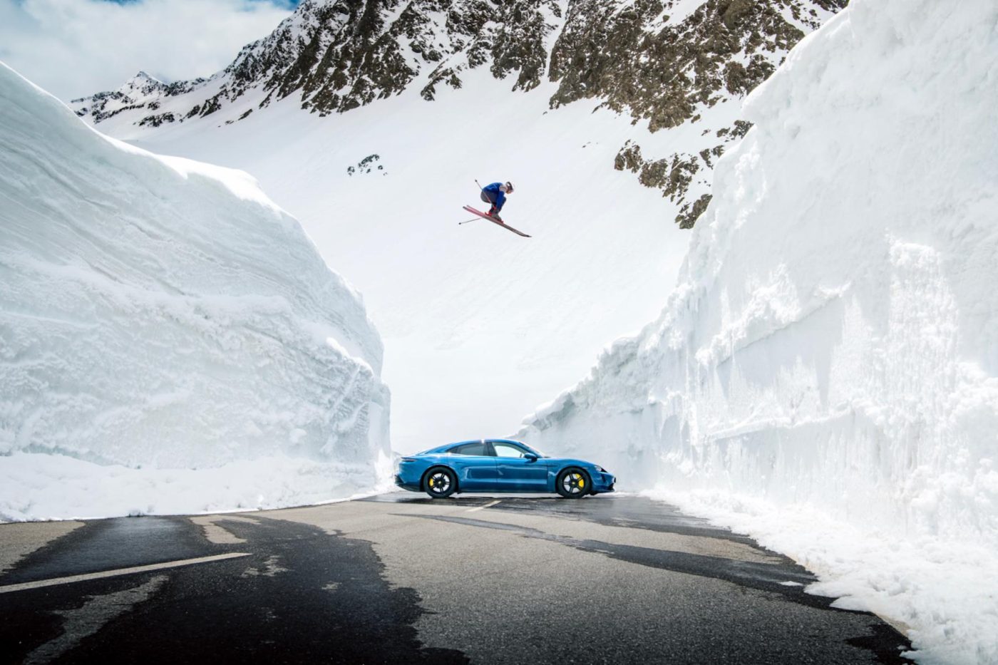 Olympic skier jumps over blue Porsche amid snow.