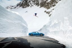 Olympic skier jumps over blue Porsche amid snow.