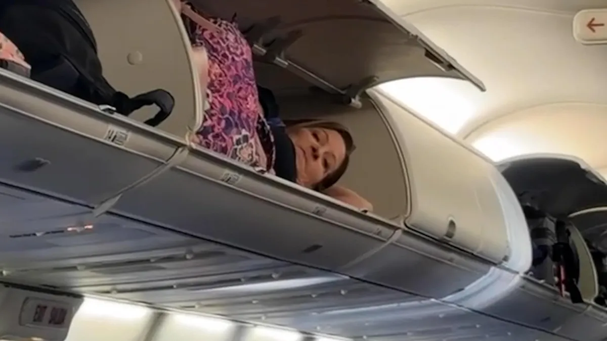 A woman lying inside an overhead locker on an airplane. 